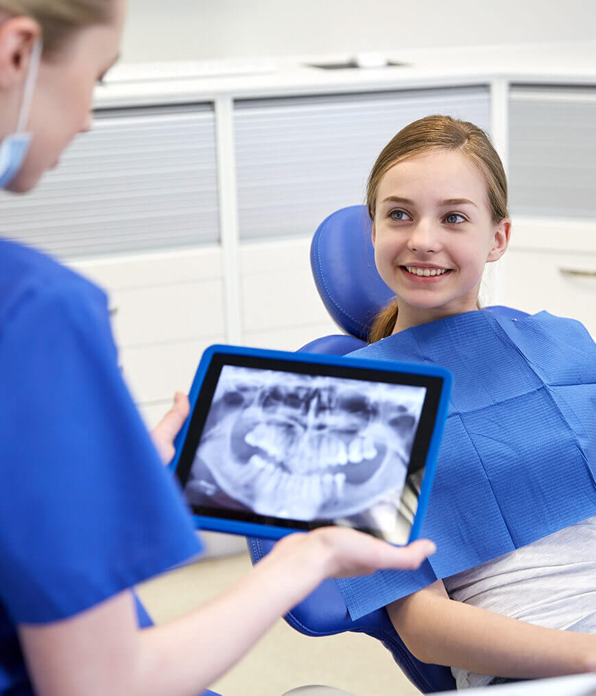 smiling girl sitting in a dental chair while her dentist examines an x-ray of her mouth