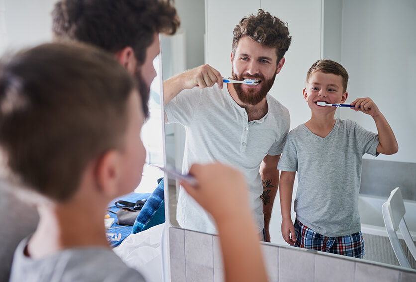 young boy and his father brushing their teeth together in front of a mirror
